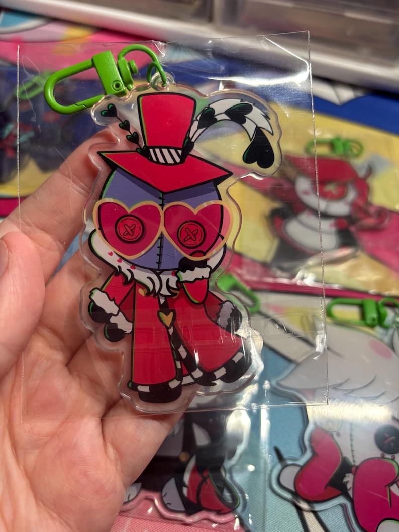 May include: A clear acrylic keychain featuring a cartoon character with a purple head, heart-shaped glasses with button-like lenses, and a red top hat. The character wears a red coat with white fur trim and has a green plastic lobster clasp attached.
