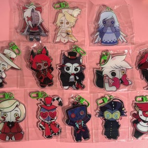 May include: A collection of thirteen individual acrylic keychains featuring stylized cartoon characters with button eyes, each enclosed in a clear plastic sleeve and attached to a bright green plastic lobster clasp hook for easy attachment.