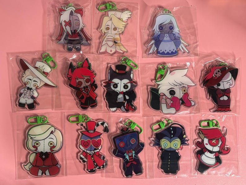 May include: A collection of thirteen individual acrylic keychains featuring stylized cartoon characters with button eyes, each enclosed in a clear plastic sleeve and attached to a bright green plastic lobster clasp hook for easy attachment.