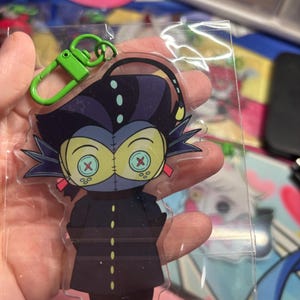 May include: A clear acrylic keychain featuring a cartoon character with dark hair, a long dark coat with yellow buttons, and large yellow eyes with red X-shaped pupils. The character has a thin antenna and a bright green plastic lobster clasp attached.