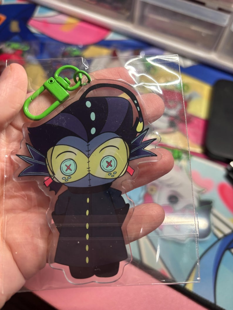 May include: A clear acrylic keychain featuring a cartoon character with dark hair, a long dark coat with yellow buttons, and large yellow eyes with red X-shaped pupils. The character has a thin antenna and a bright green plastic lobster clasp attached.