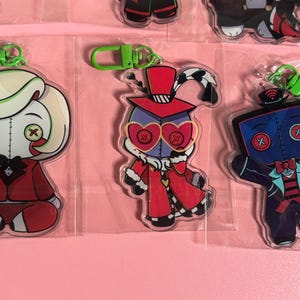 May include: Three acrylic keychains featuring stylized cartoon characters with button eyes. The left character has blonde hair and a red suit. The center character wears a red top hat and heart-shaped glasses. The right character is blue with a bowtie.