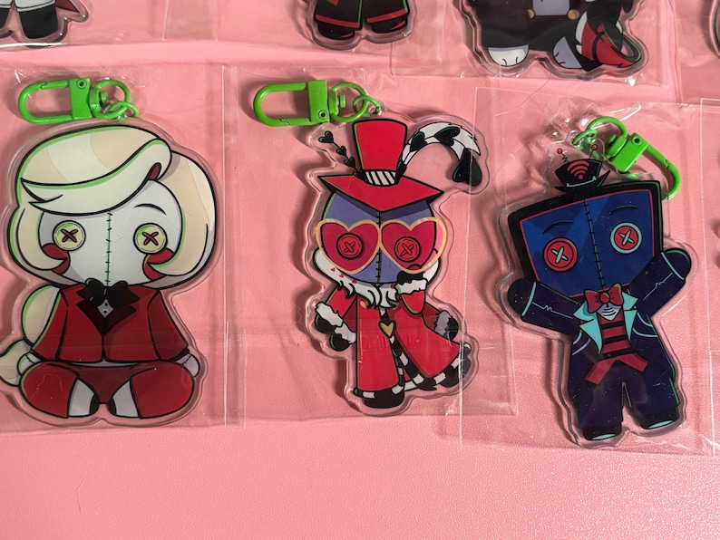 May include: Three acrylic keychains featuring stylized cartoon characters with button eyes. The left character has blonde hair and a red suit. The center character wears a red top hat and heart-shaped glasses. The right character is blue with a bowtie.