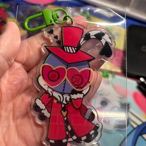 May include: A clear acrylic keychain featuring a cartoon character with a purple head, heart-shaped glasses with button details, and a red coat with white fur trim. The character wears a red top hat with a feather and has a bright green plastic clip.