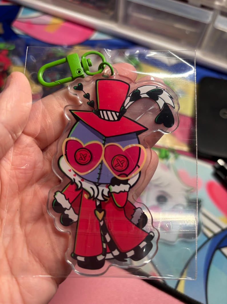 May include: A clear acrylic keychain featuring a cartoon character with a purple head, heart-shaped glasses with button details, and a red coat with white fur trim. The character wears a red top hat with a feather and has a bright green plastic clip.