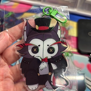 May include: A clear acrylic keychain featuring a stylized cartoon character with dark purple fur, button eyes, and a stitched face. The character wears a black top hat with a red band, a red bowtie, and a black suit. A bright green plastic lobster clasp is attached.