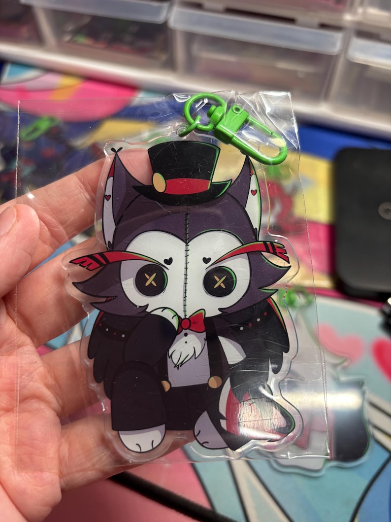 May include: A clear acrylic keychain featuring a stylized cartoon character with dark purple fur, button eyes, and a stitched face. The character wears a black top hat with a red band, a red bowtie, and a black suit. A bright green plastic lobster clasp is attached.