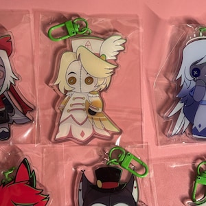 May include: A collection of acrylic character keychains in clear plastic packaging, each featuring a green lobster clasp. The designs include stylized figures with button eyes, including a red and white character, a blonde figure, and a blue angel.