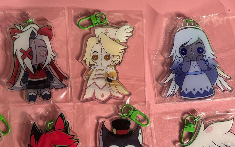 May include: A collection of acrylic character keychains in clear plastic packaging, each featuring a green lobster clasp. The designs include stylized figures with button eyes, including a red and white character, a blonde figure, and a blue angel.
