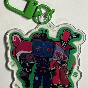 May include: A clear acrylic keychain featuring a green lobster clasp and a printed illustration of three stylized characters with button eyes. The central figure has a blue square head, while the flanking figures wear pink and red outfits.