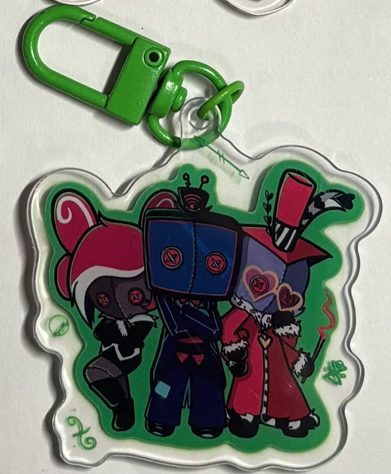 May include: A clear acrylic keychain featuring a green lobster clasp and a printed illustration of three stylized characters with button eyes. The central figure has a blue square head, while the flanking figures wear pink and red outfits.
