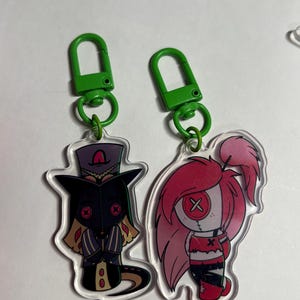 May include: Two acrylic keychains with green plastic lobster clasps. One features a character in a purple top hat and black mask with red button eyes. The other depicts a character with long pink hair and a white face with a red button eye.