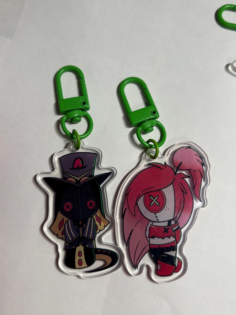 May include: Two acrylic keychains with green plastic lobster clasps. One features a character in a purple top hat and black mask with red button eyes. The other depicts a character with long pink hair and a white face with a red button eye.
