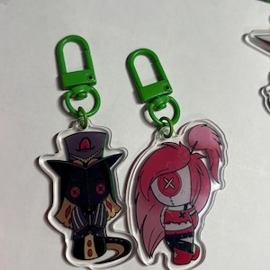 May include: Two acrylic keychains with green plastic clasps. One features a character in a dark purple top hat and coat with red button eyes. The other depicts a character with long pink hair, a red top, and a single large button eye with an X.