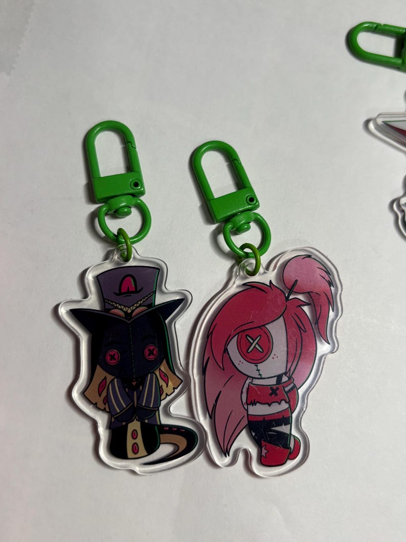 May include: Two acrylic keychains with green plastic clasps. One features a character in a dark purple top hat and coat with red button eyes. The other depicts a character with long pink hair, a red top, and a single large button eye with an X.