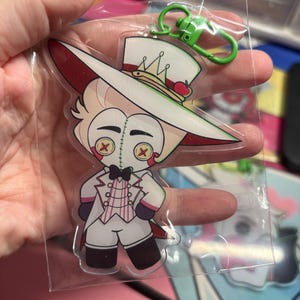 May include: A clear acrylic keychain featuring a cartoon character with blonde hair, a white top hat with a small red apple, and a white suit with a pink striped vest. The character has button eyes with red X stitching and a black bowtie.