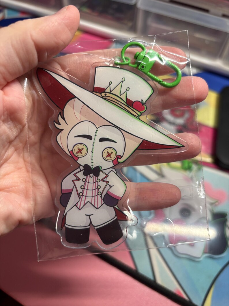 May include: A clear acrylic keychain featuring a cartoon character with blonde hair, a white top hat with a small red apple, and a white suit with a pink striped vest. The character has button eyes with red X stitching and a black bowtie.