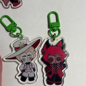 May include: Two acrylic keychains featuring cartoon characters with button eyes. One character wears a white suit and top hat, while the other has red hair and a red suit. Both are attached to bright green plastic lobster claw clasps.