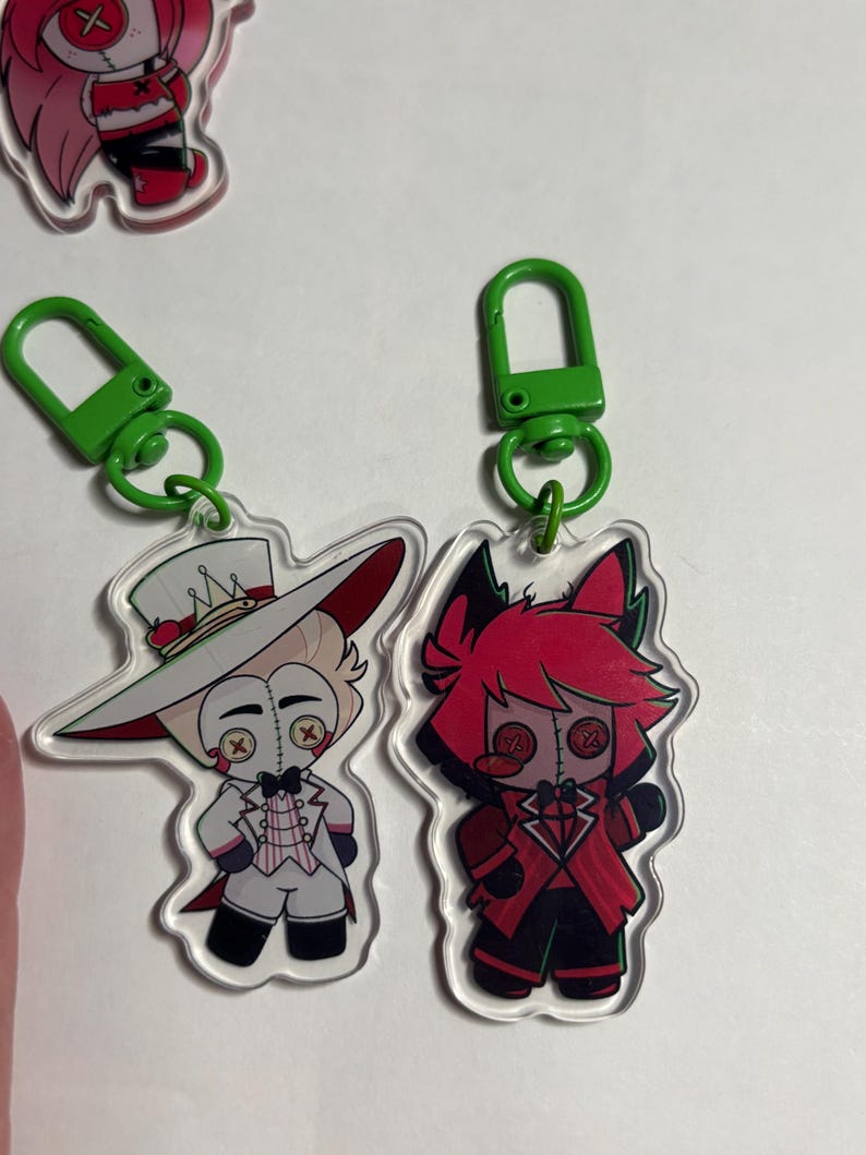 May include: Two acrylic keychains featuring cartoon characters with button eyes. One character wears a white suit and top hat, while the other has red hair and a red suit. Both are attached to bright green plastic lobster claw clasps.