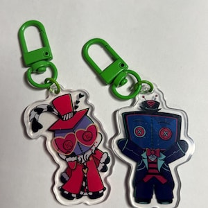 May include: Two acrylic keychains with bright green plastic clasps. The left keychain features a character in a red coat and top hat with heart-shaped glasses. The right keychain depicts a blue-faced character with a television-shaped head.