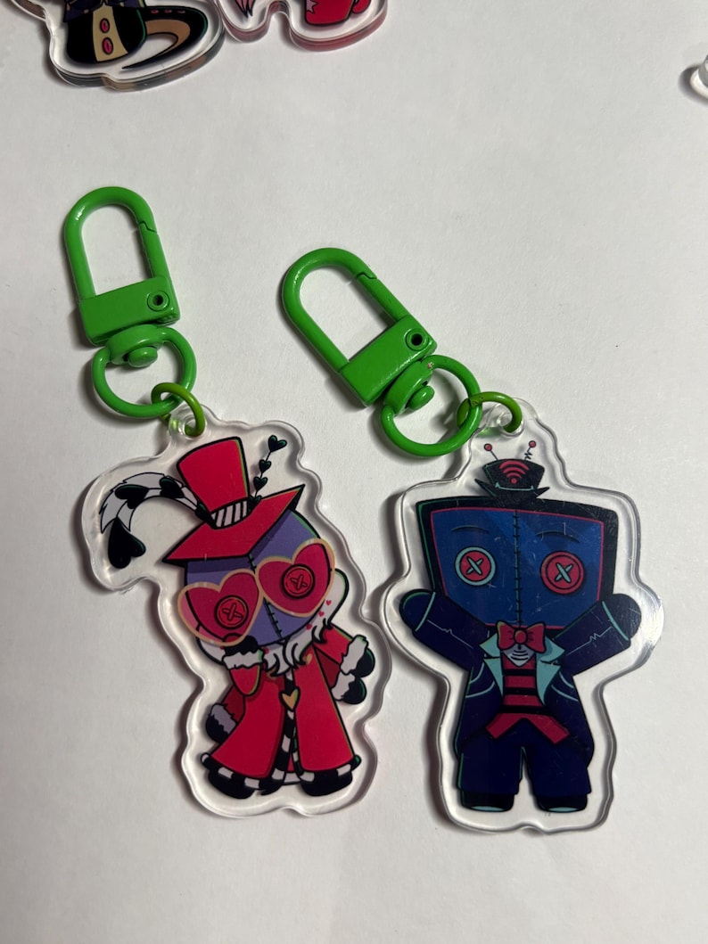 May include: Two acrylic keychains with bright green plastic clasps. The left keychain features a character in a red coat and top hat with heart-shaped glasses. The right keychain depicts a blue-faced character with a television-shaped head.
