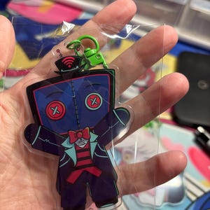 May include: A clear acrylic keychain featuring a cartoon character with a dark blue square head, red button eyes, and a stitched seam down the center. The character wears a black top hat with a red Wi-Fi symbol, a red bowtie, and a dark suit.