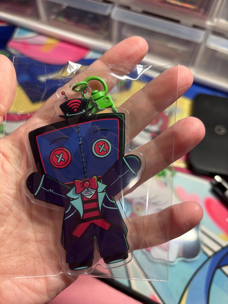 May include: A clear acrylic keychain featuring a cartoon character with a dark blue square head, red button eyes, and a stitched seam down the center. The character wears a black top hat with a red Wi-Fi symbol, a red bowtie, and a dark suit.