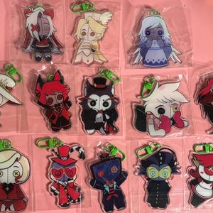 May include: A collection of thirteen acrylic keychains featuring stylized cartoon characters with button eyes, each individually packaged in a clear plastic sleeve and attached to a bright green plastic lobster clasp hook for easy attachment.