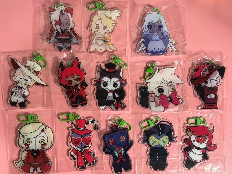 May include: A collection of thirteen acrylic keychains featuring stylized cartoon characters with button eyes, each individually packaged in a clear plastic sleeve and attached to a bright green plastic lobster clasp hook for easy attachment.
