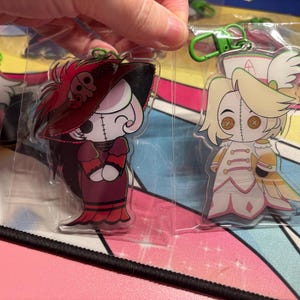 May include: Two clear acrylic keychains featuring cartoon characters with button eyes and stitched faces. One character wears a dark red dress and wide-brimmed hat, while the other wears a white and gold outfit with a nurse-style cap and green clip.