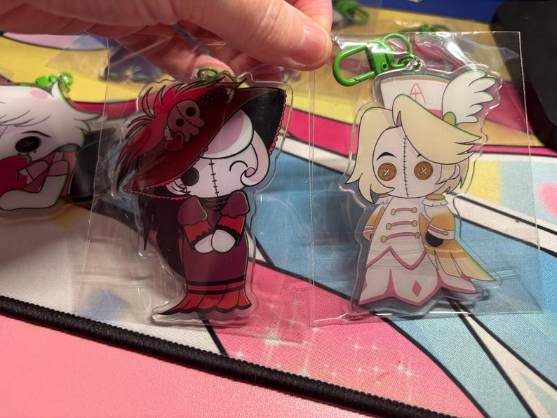 May include: Two clear acrylic keychains featuring cartoon characters with button eyes and stitched faces. One character wears a dark red dress and wide-brimmed hat, while the other wears a white and gold outfit with a nurse-style cap and green clip.