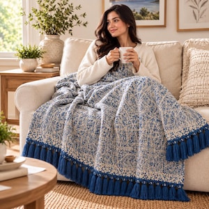 May include: A woven throw blanket featuring a blue and cream textured pattern with a repeating geometric design. The blanket is finished with long, thick blue tassels along the bottom edge, providing a decorative border to the fabric.