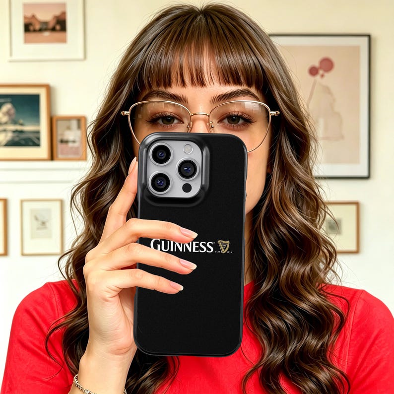 May include: Black phone case with the Guinness logo in white. The case has a glossy finish and a camera cutout. The phone case is held in front of a person's face.