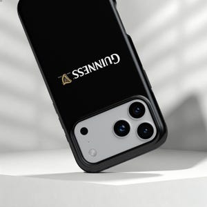 May include: Black phone case with the Guinness logo in white and gold. The case has a modern design with a black border and a silver camera module.