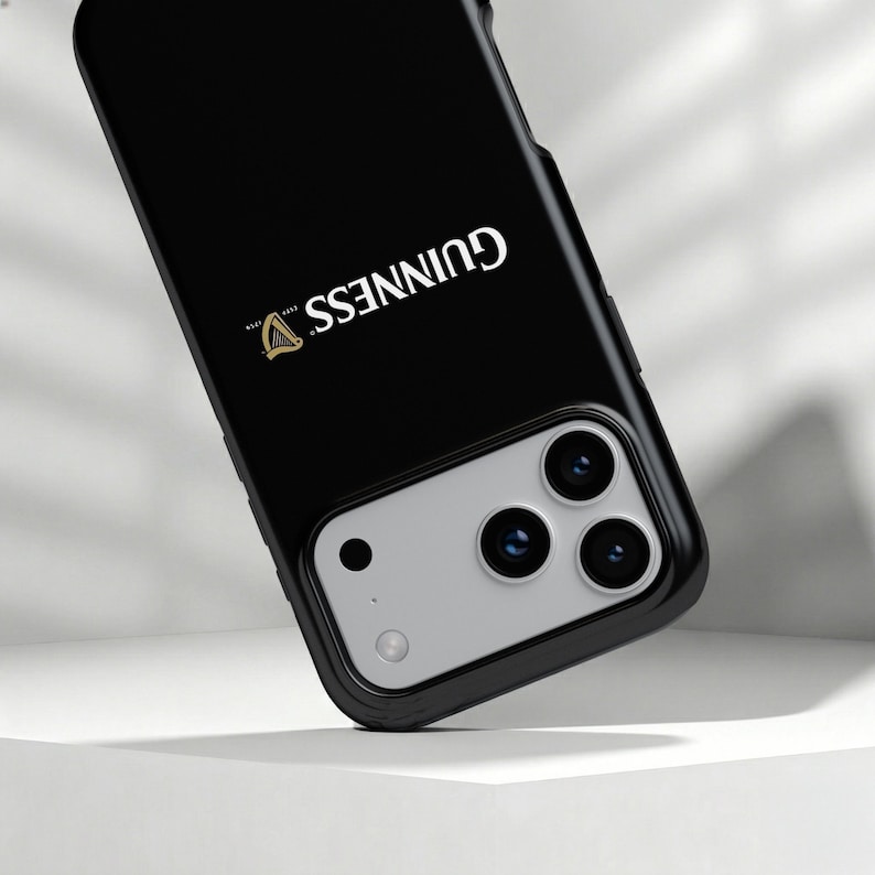 May include: Black phone case with the Guinness logo in white and gold. The case has a modern design with a black border and a silver camera module.
