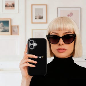 May include: Black phone case featuring the Guinness logo. The case is held by a person wearing black sunglasses and a black turtleneck. Framed artwork is visible in the background.
