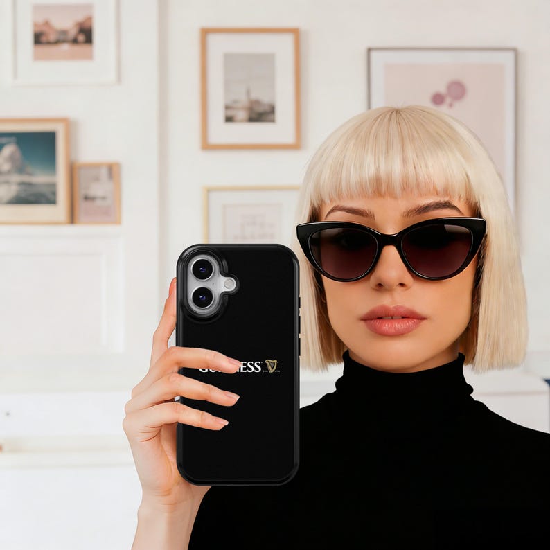 May include: Black phone case featuring the Guinness logo. The case is held by a person wearing black sunglasses and a black turtleneck. Framed artwork is visible in the background.