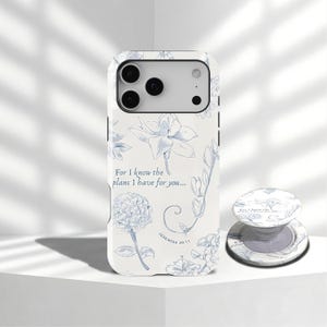 Op de afbeelding: Witte telefoonhoes met een blauw bloemenpatroon en de tekst "For I know the plans I have for you... Jeremiah 29:11." Een bijpassende telefoonhouder is ook zichtbaar. De hoes heeft een camera-uitsparing en een gladde afwerking.