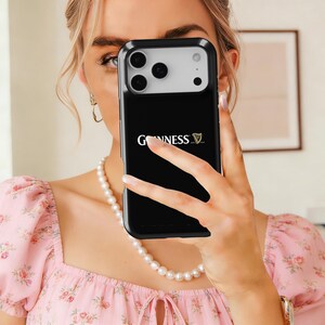May include: Black phone case featuring the Guinness logo in white. The case is held in front of a person's face. The phone case has a black border and a silver camera section. The person is wearing a pink floral top and a pearl necklace.