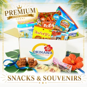 Può includere: Una scatola di cartone riempita con vari snack e souvenir giapponesi di Okinawa. La scatola presenta il logo Okinawa Discovery Shop con un fiore di ibisco rosso. Sono inclusi pacchetti di snack colorati, due piccole statue shisa marroni e un fiore di ibisco.