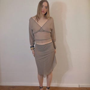 Könnte beinhalten: Ein zweiteiliges Outfit in transparentem Taupe. Das Oberteil hat einen V-Ausschnitt und lange Ärmel mit dunklem Besatz. Der Rock ist drapiert und asymmetrisch. Das Outfit wird mit Leopardenmuster-Absätzen kombiniert.