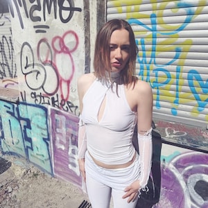 May include: A white halter top with a keyhole cutout, paired with matching white trousers. The top features sheer, long sleeves. The background includes graffiti art on a wall and a metal shutter.