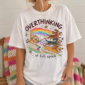 Puede incluir: Una camiseta blanca de manga corta con un gráfico colorido de un gato naranja en patines siendo arrastrado por tres perros salchicha marrones. El texto dice OVERTHINKING at full speed, junto a un arcoíris, estrellas y pequeños dibujos.