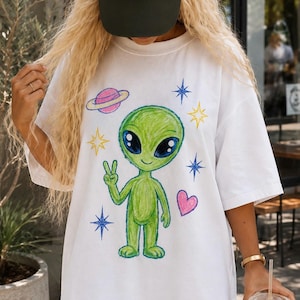 Op de afbeelding: Een wit T-shirt met korte mouwen en een kleurrijke tekening in kleurpotloodstijl van een groene alien die een vredesteken maakt. De afbeelding bevat een roze planeet met een ring, een roze hart en diverse blauwe en gele vierpuntige sterren.