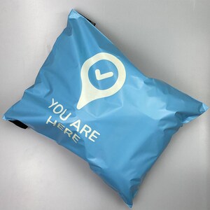 May include: A light blue shipping bag with the words "YOU ARE HERE" in white text. A white location pin graphic with a checkmark is above the text. The bag is sealed and appears to be empty.