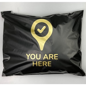 Könnte beinhalten: Schwarzer Poly-Versandbeutel mit goldener Standortmarkierung und dem Schriftzug "YOU ARE HERE". Die Standortmarkierung hat ein Häkchen. Der Beutel ist versiegelt und versandfertig.