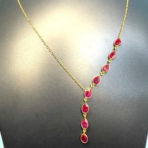May include: A gold-colored necklace featuring a series of teardrop-shaped, red gemstones. The gemstones are set in individual gold bezels and cascade down the center of the necklace. The chain is delicate and also gold-colored.