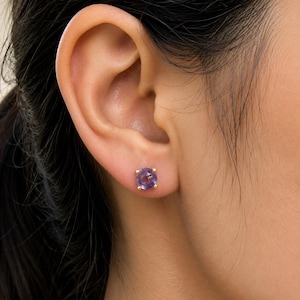 May include: A round, purple gemstone stud earring set in a four-prong gold metal mount. The earring is positioned in the earlobe, featuring a faceted surface that reflects light, showcasing a deep violet hue and a polished metallic gold setting.