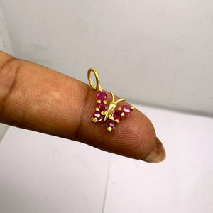 Puede incluir: Un colgante de mariposa dorado con piedras preciosas de color rubí. Las alas están adornadas con piedras redondas de color rojo vibrante, y el cuerpo y las antenas son de oro. El colgante está diseñado para ser usado como joya.