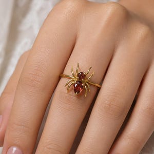 Könnte beinhalten: Ein goldener Ring mit einem Spinnenmotiv, dessen Körper aus zwei tiefroten, birnenförmigen Edelsteinen besteht. Die Spinne hat acht schlanke goldene Beine, die von den zentralen Steinen ausgehen, welche vertikal auf dem schmalen Goldring sitzen.
